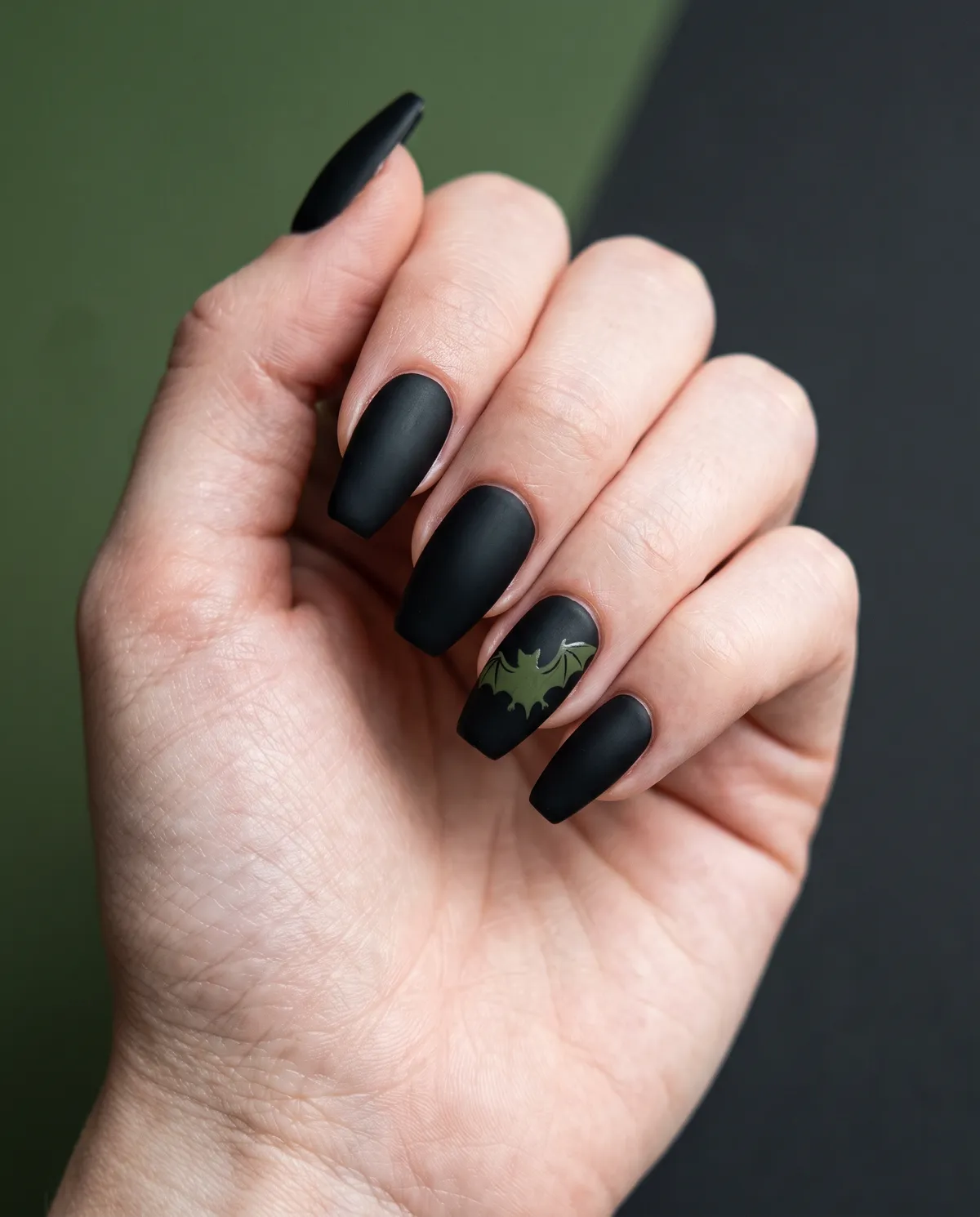 Bat Wing Matte — coffin halloween nails with matte black gel base on coffin nails with a hand-painted olive-green bat silhouette on the ring…